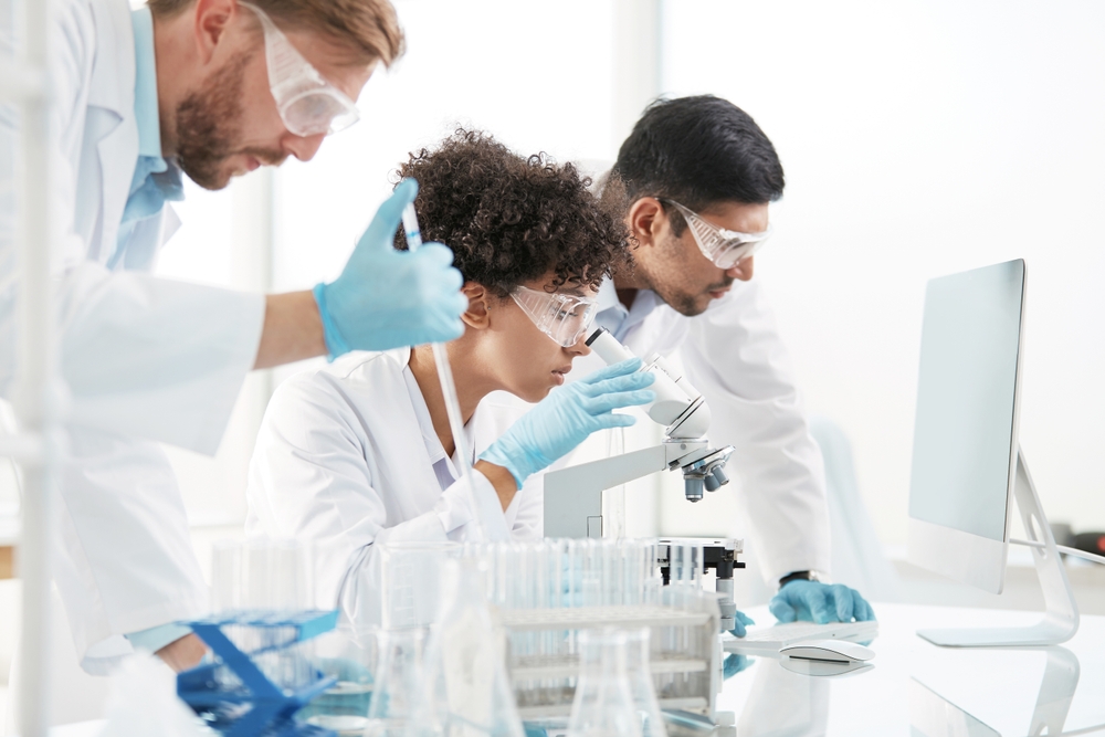 Três pessoas em ambiente de laboratório utilizam equipamento científico: uma observa uma amostra ao microscópio, enquanto as outras acompanham a análise junto a uma bancada com tubos de ensaio, frascos e um computador, todas com batas, luvas e óculos de proteção.