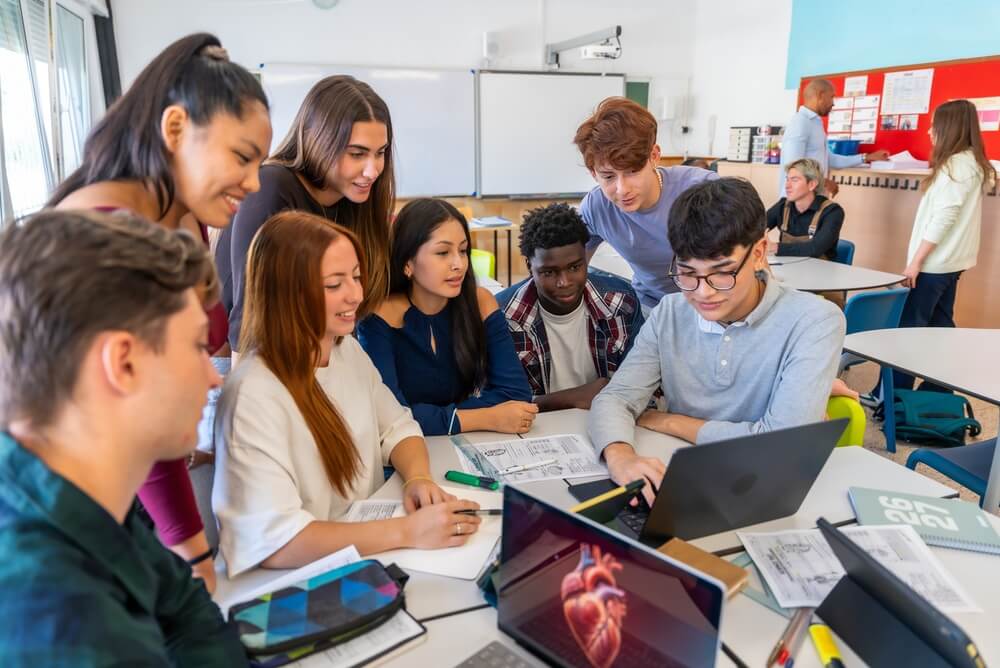 Grupo de alunos numa sala de aula, a trabalhar em conjunto num computador portátil e a consultar materiais de estudo.