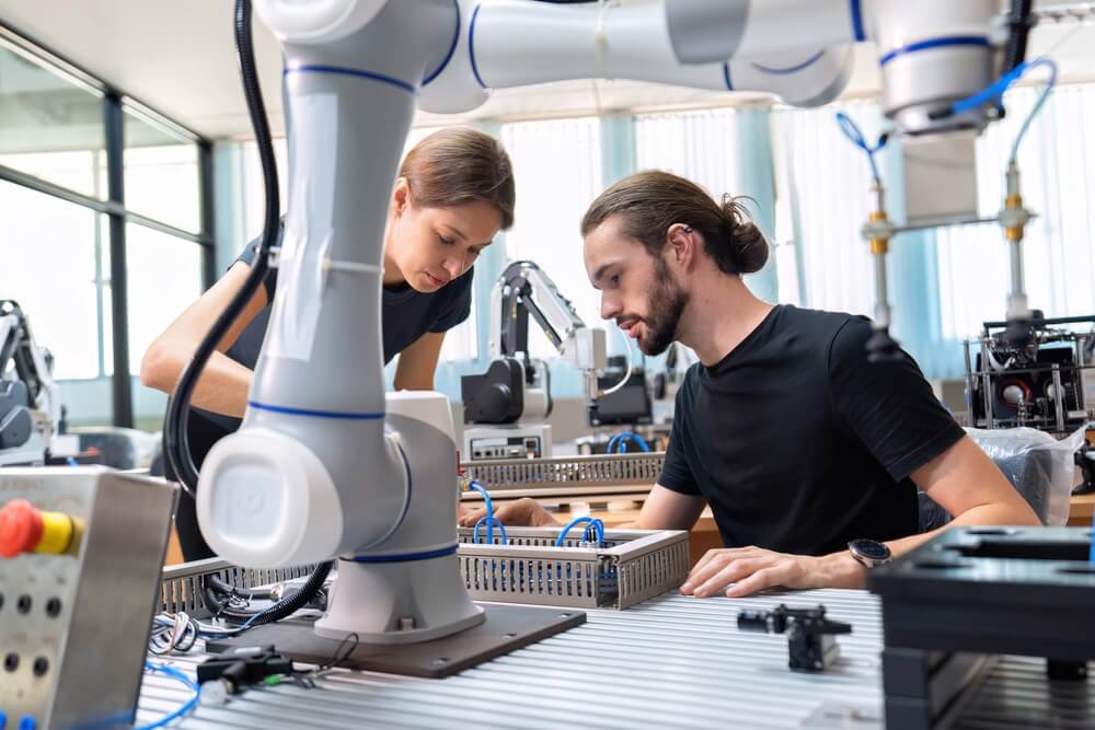 Dois profissionais trabalham com um braço robótico numa bancada de laboratório dedicada à robótica e à automação industrial.
