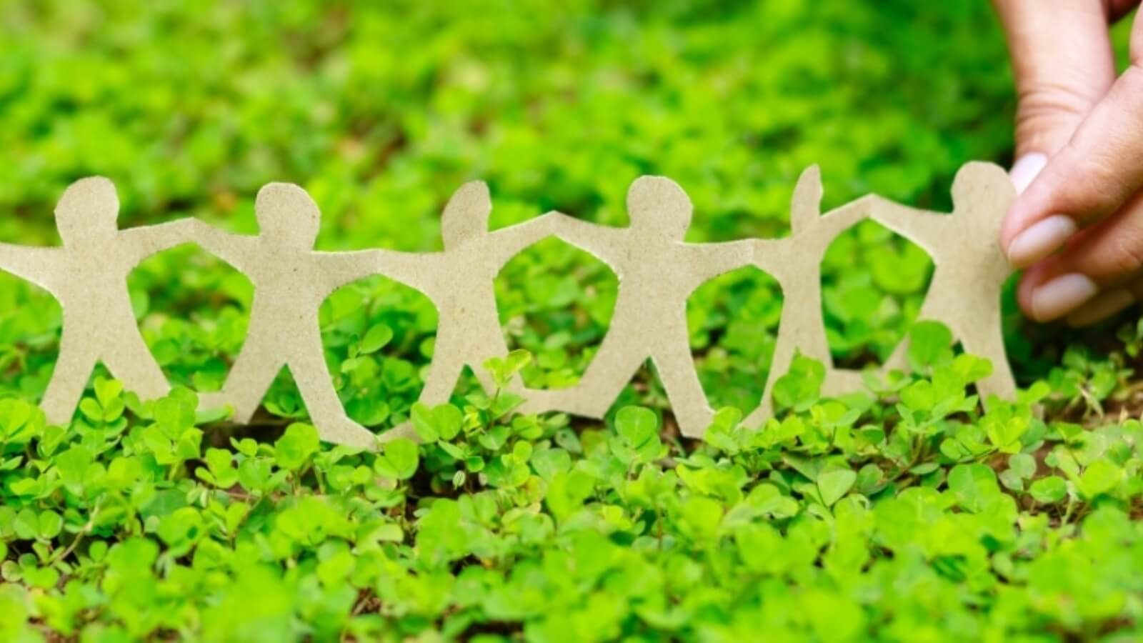 Mão a segurar uma corrente de figuras humanas recortadas em cartão, sobre um fundo de vegetação verde.